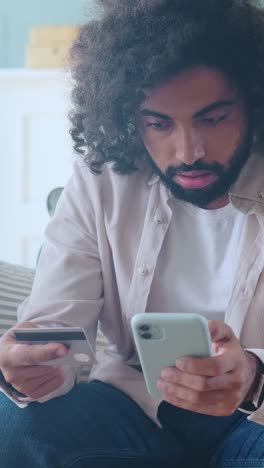 man checks his phone while holding a credit card in a cozy living room setting