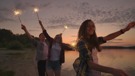 Alegres-Amigos,-Hombres-Y-Mujeres,-Corren-Por-La-Playa-Al-Atardecer-Sosteniendo-Brillantes-Fuegos-Artificiales-Y-Luces-Desbocadas-En-Cámara-Lenta.-Baile-Y-Fiesta-Al-Atardecer-En-La-Playa