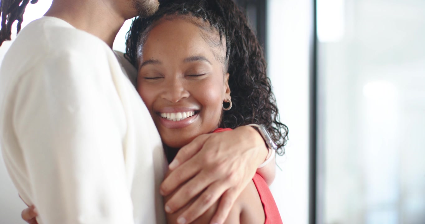 African American couple sharing embrace after man smiling down, comforting by window with red top