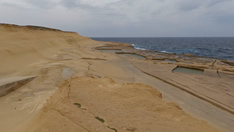 Vista-Aérea-Sobre-Las-Plantas-De-Sal-De-Malta-O-Las-Bandejas-De-Evaporación-De-Sal-En-La-Costa-Rocosa-De-Malta-Con-Un-Paisaje-Seco-Con-Vistas-Al-Mar-Con-Olas-Tranquilas-En-Un-Día-Nublado