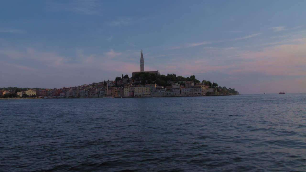 Boat view of Rovinj old town skyline and church tower at dusk on the Adriatic Sea, Croatia. Calm water and pastel twilight sky create a serene mood