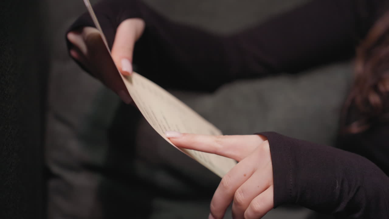 Close up view of woman holding menu and pointing at specific item with finger while seated in soft green booth. Shot emphasizes hands, gesture, and engagement with content in relaxed setting