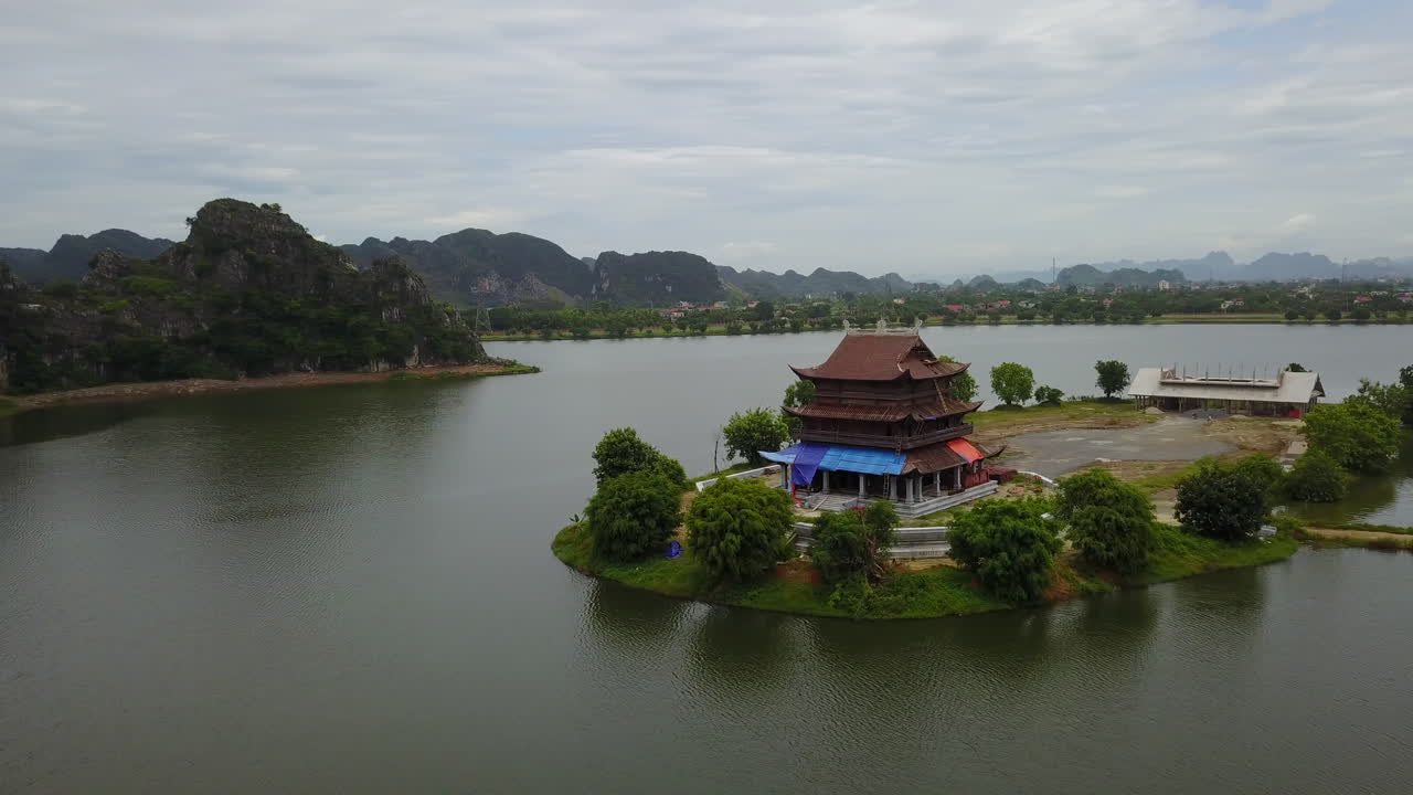 Experience a captivating aerial orbit around the serene Dinh Thanh Cao Son temple in Trang An, Ninh Binh, Vietnam, beautifully situated by the water's edge amidst the region's striking karst landscape