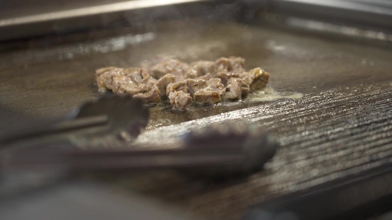 Sottili pezzi di pollo sfrigolano nell'olio su una piastra metallica calda, mentre delle pinze in acciaio inossidabile riposano in primo piano.