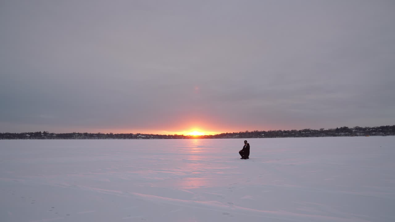 Man stands alone on a vast frozen lake during sunset, with the golden light reflecting on the icy surface and a winter sky overhad