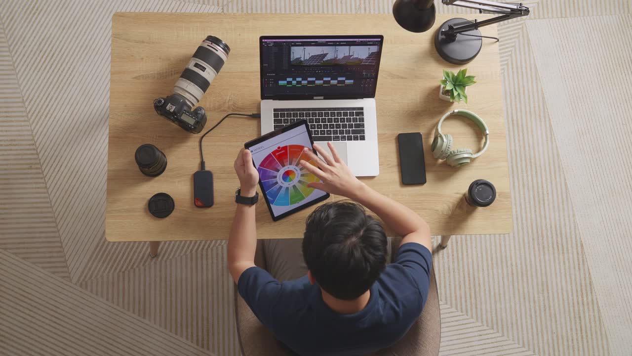 vista superior de un editor masculino mirando los esquemas de colores en una tableta mientras está sentado en el espacio de trabajo usando una computadora portátil al lado de la cámara editando el video en casa