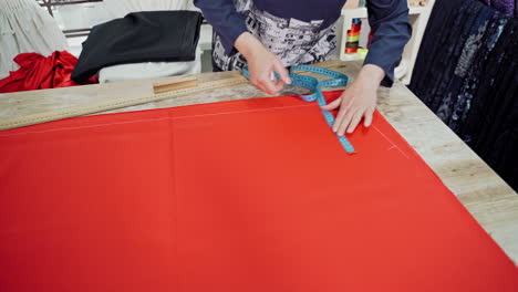 woman's hands measuring red fabric with blue tape measure on a table. seamstress works with red cloth sketching lines with a chalk. sewing and handicraft concept.