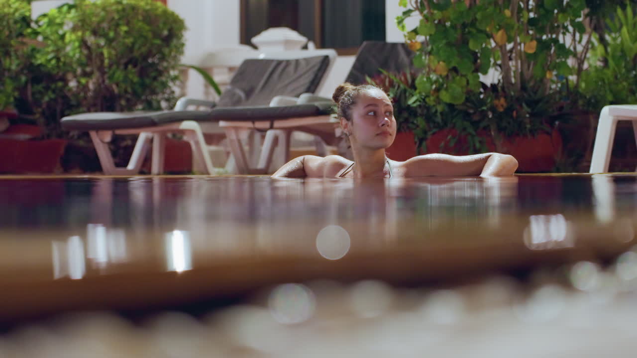 Donna che si rilassa in una piscina di un resort sull'isola, appoggiata al bordo vicino a sedie a sdraio e piante tropicali, bokeh in primo piano con messa a fuoco superficiale, luce calda del tardo pomeriggio, atmosfera contemplativa, leggere increspature e spa.