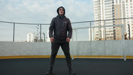 sportsman performing deep squats with hands clasped, focusing on preparation for exercise on outdoor sports ground, soccer ball in foreground, surrounded by urban high buildings and goal post