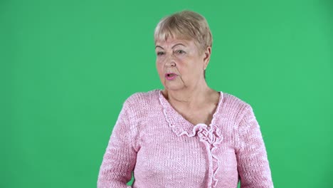 portrait elderly woman shaking his head its not me, i dont know whats going on. gray haired grandmother with short hair in a pink sweater on green screen at studio. close up