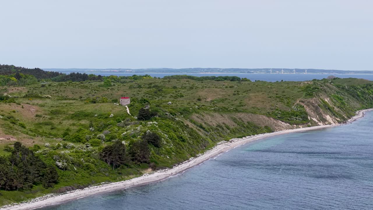 Drone footage showcasing a scenic coastal peninsula in Denmark with green vegetation, shrubs and a calm blue sea along a curving shoreline under a clear sky