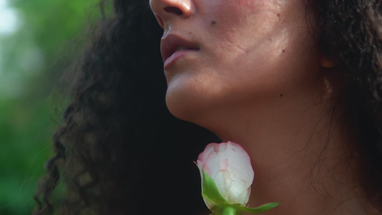 Close-up on a woman's lower face and curly hair, with a white rosebud held at her neck. Background is softly blurred greenery