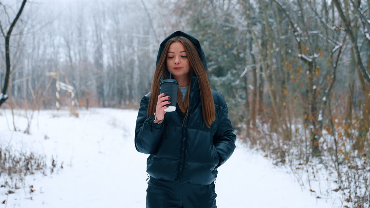 Young woman wearing black jacket with a hood goes by the winter park. Happy lady walks drinking coffee and smiling.