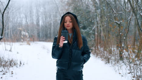 young woman wearing black jacket with a hood goes by the winter park. happy lady walks drinking coffee and smiling.