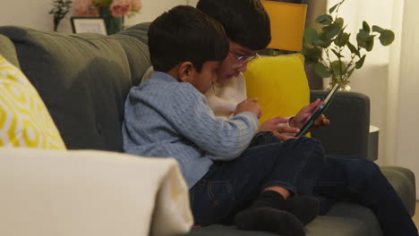 two young boys sitting on sofa at home playing games or streaming onto digital tablet together 4