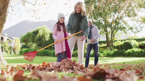 video of happy caucasian grandparents and granddaughter raking up autumn leaves in sunny garden