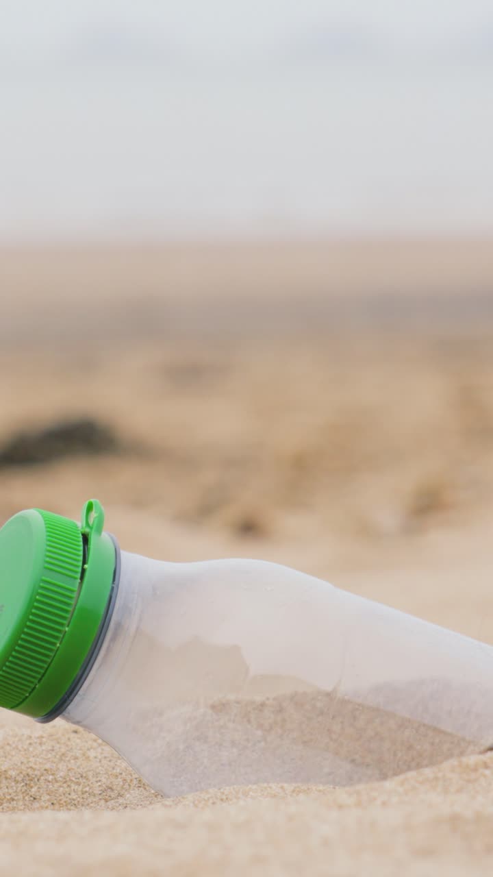 Vertical Video Reveal of Plastic Drink Bottle Sticking Out of Soft Sand on Beach with Shallow Depth of Field. Pollution Concept Causing Hazards to Natural Environment