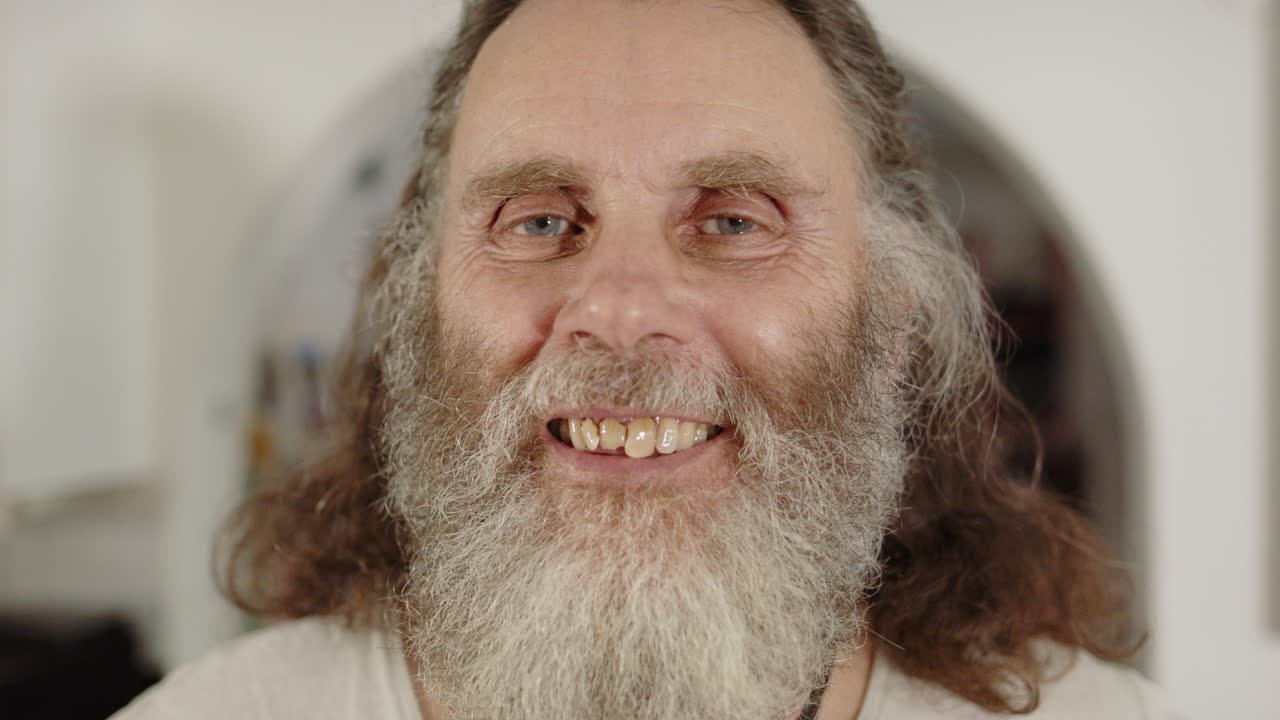 Unkempt senior man with lots of facial hair smiling as he looks into camera lens