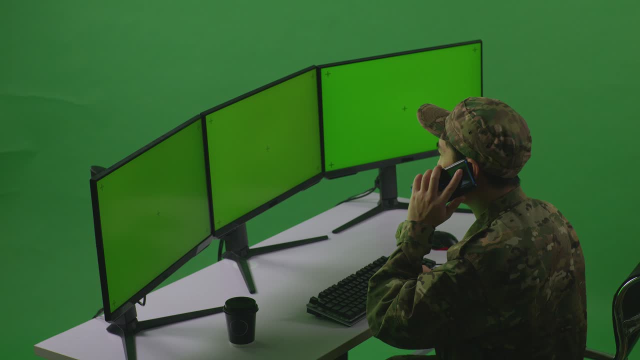 vista lateral de un soldado asiático hablando en teléfono móvil mientras trabaja con un simulacro de varios monitores de computadora en un estudio de pantalla verde