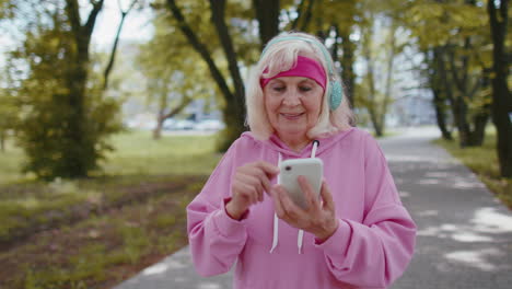 Athletic-senior-sport-runner-woman-wearing-earphones-listening-music-from-smartphone-in-morning-park