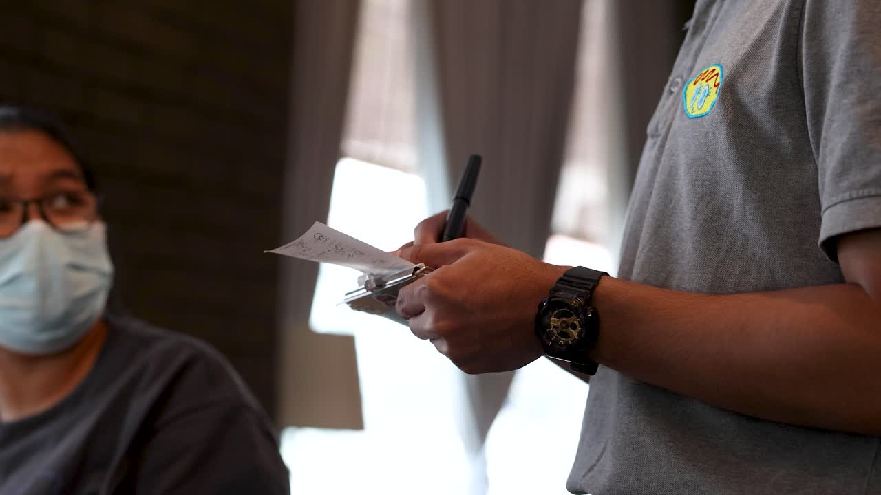 Waiter writes order on clipboard beside masked diner in softly lit, casual dining environment