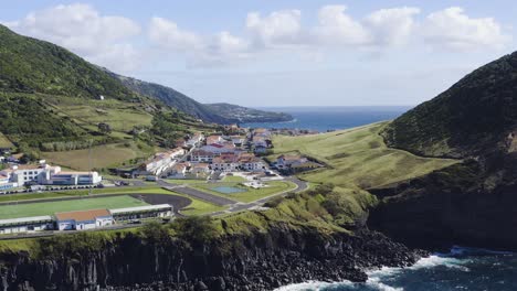 Velas,-Sao-Jorge-island,-Azores,-Portugal-Aerial-view-of-whitewashed-houses-village,-skate-park,-sports-field,-with-surrounding-mountains,-vegetation-and-lava-formations-seaside-drone-footage