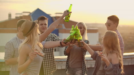 russian people clinks glasses and drinks beer from green bottels on the party with friends on the roof. they eat hot pizza after.