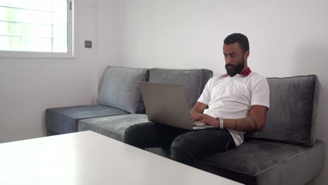 hombre molesto trabajando y escribiendo en su computadora portátil mientras está sentado en el sofá