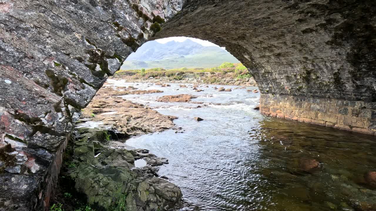 Camera smoothly pans beneath an old stone bridge, revealing a flowing river, rocky banks, and distant mountains in natural daylight on the Isle of Skye