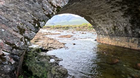 camera smoothly pans beneath an old stone bridge, revealing a flowing river, rocky banks, and distant mountains in natural daylight on the isle of skye