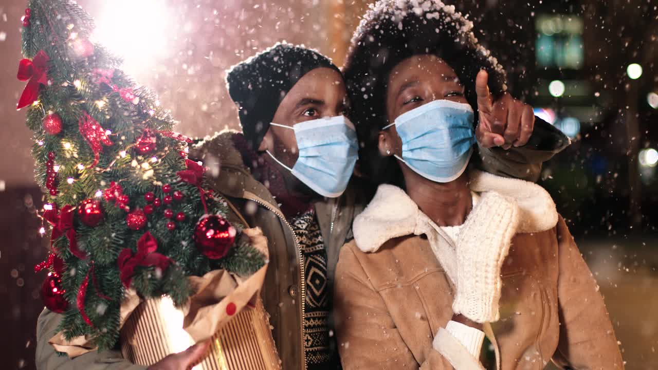 크리스마스에 거리에 눈이 내리는 동안 얼굴 마스크를 쓰고 이야기하고 둘러보는 즐거운 아프리카계 미국인 커플의 클로즈업 보기