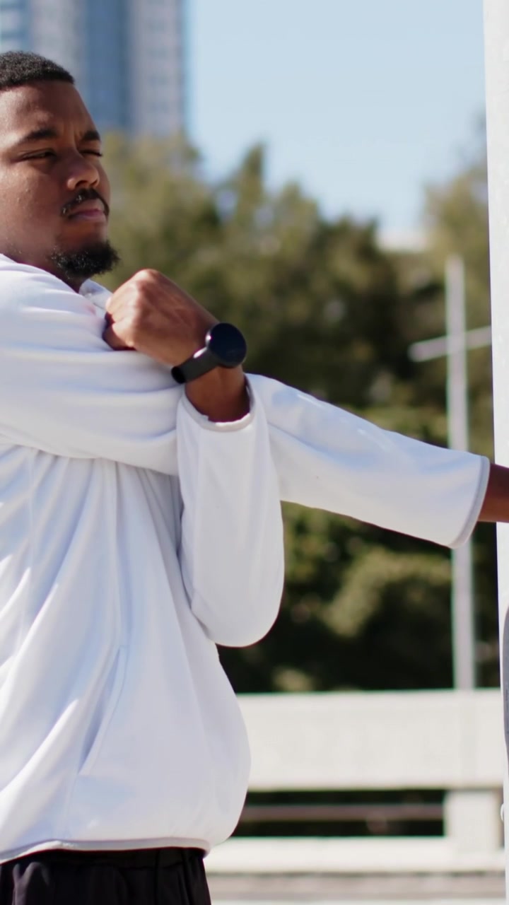 Verticale video: Man tilt rechterarm op en rekt schouder op dakterras, draagt hoodie en smartwatch.