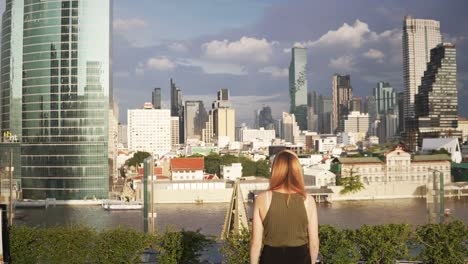 a young woman squats on a hotel roof, gets up, and walks toward the bangkok city skyline