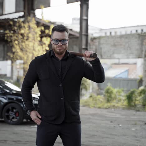 strong man with a bat in abandoned place. stylish man in glasses and black wearing moving forward on expensive car background inside the old empty plant.