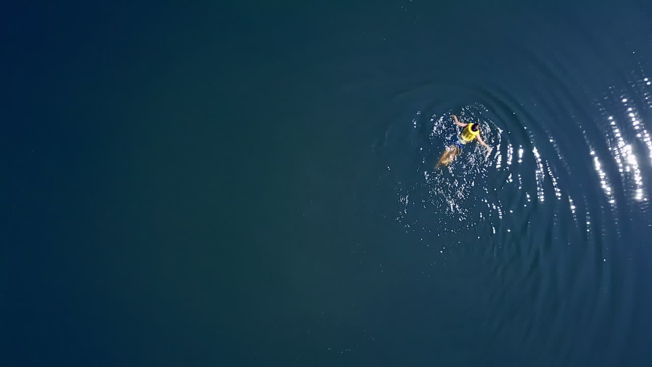 Little boy is swimming in a clear blue water in a sunny day in summer. Teenage boy wearing yellow life vest bathing alone on the water backgound. Aerial view