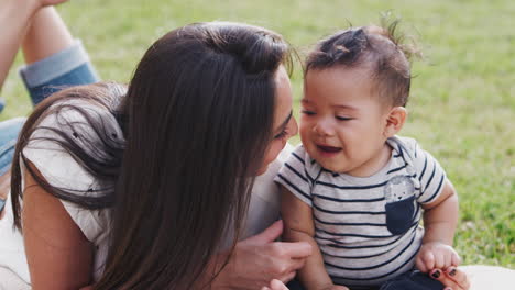 millennial hispanic mother lying on the grass in the park playing with her baby boy, close up