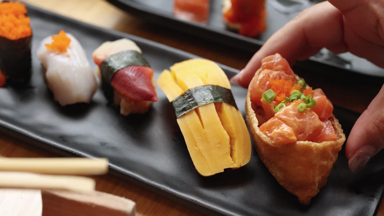 Hand reaches for fresh assorted nigiri sushi on black plate, natural lighting, close-up view