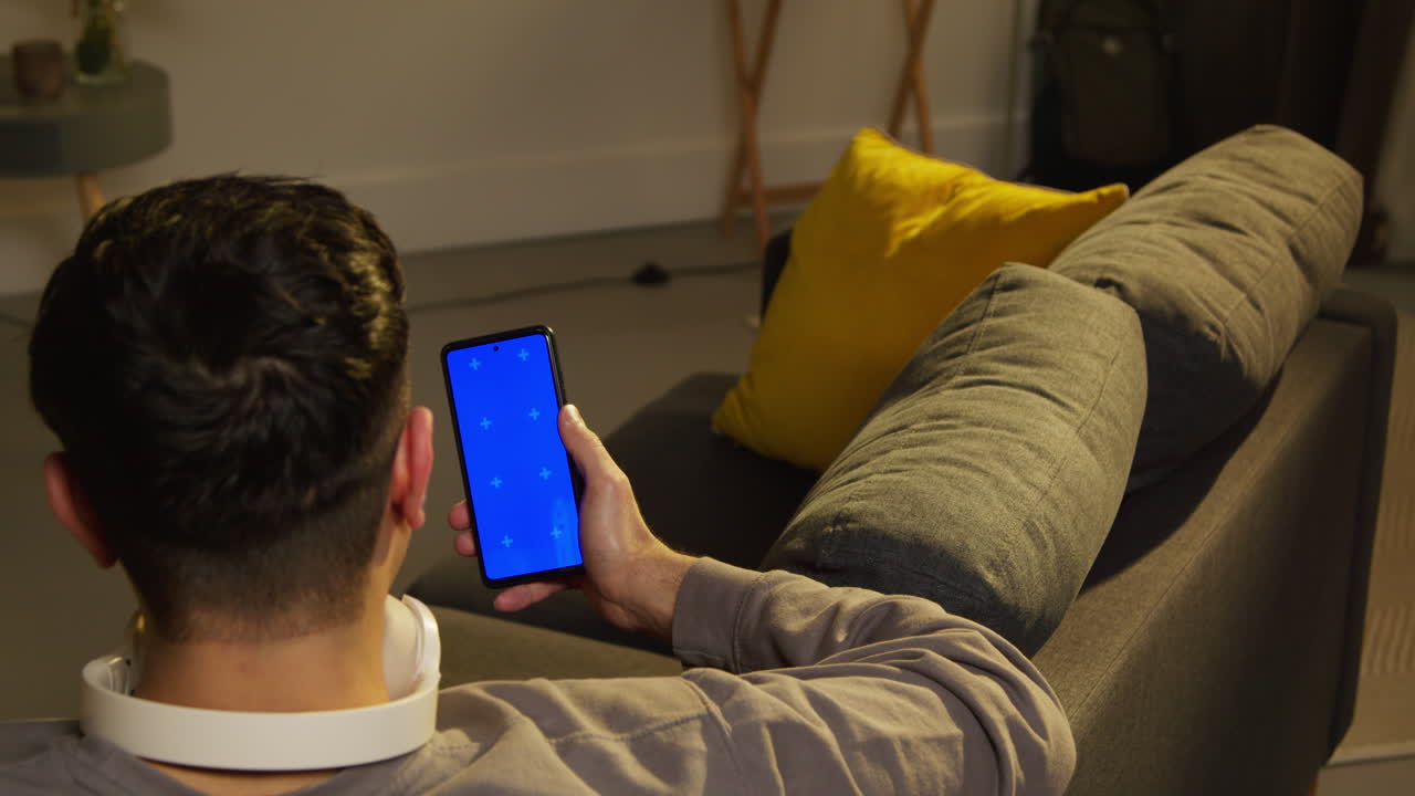 vista trasera de un hombre pasando la noche en casa sentado en el sofá mirando la pantalla azul del teléfono móvil