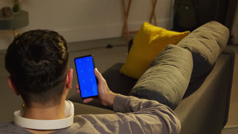 rear view of man spending evening at home sitting on sofa looking at blue screen mobile phone