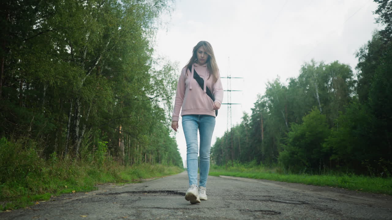 Confident woman in pink hoodie and jeans walking along tarred countryside road with trees on both sides, natural light casting soft shadows, peaceful mood captured in motion from low front angle
