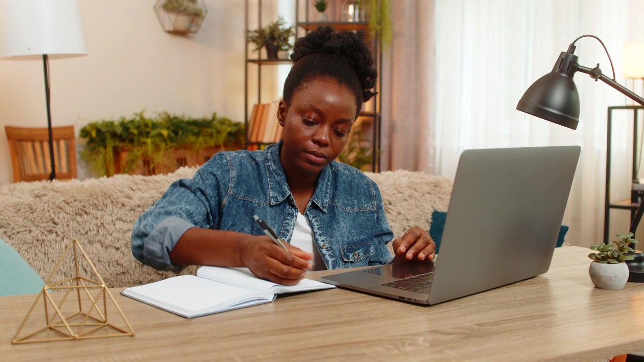 African american young woman studies online with laptop and notebook takes write notes at home table