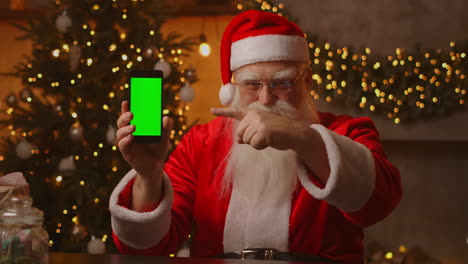 santa points his finger at the green screen mobile phone screen. christmas sale. elderly santa claus shows a screen with a chromakey. high quality 4k footage