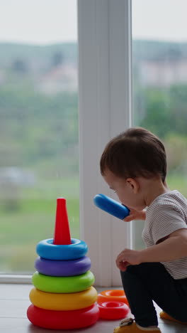 toddler boy comes up to a colorful pyramid near the panoramic window. active happy kid assembles the toy. vertical video