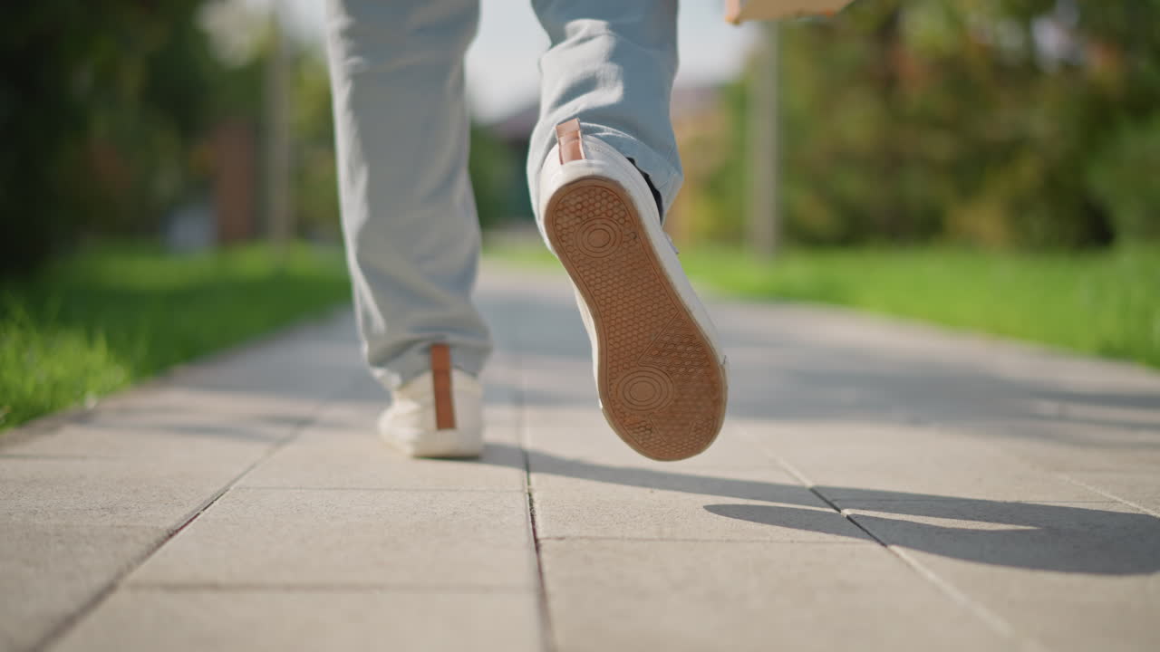 closeup feet walking on sidewalk path, person in light jeans carrying sketchbook, strolling through sunlit urban park lane toward outdoor painting spot focus on sneaker soles, pavement texture, soft