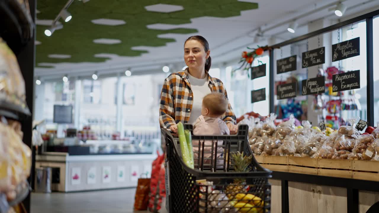 A happy brunette girl in a plaid shirt pushes a cart along with her little child and groceries during her shopping in a supermarket