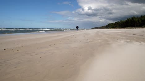 Día-Ventoso-En-Una-Playa-De-Arena,-Arena-Que-Sopla,-Hombre-Caminando-En-La-Distancia