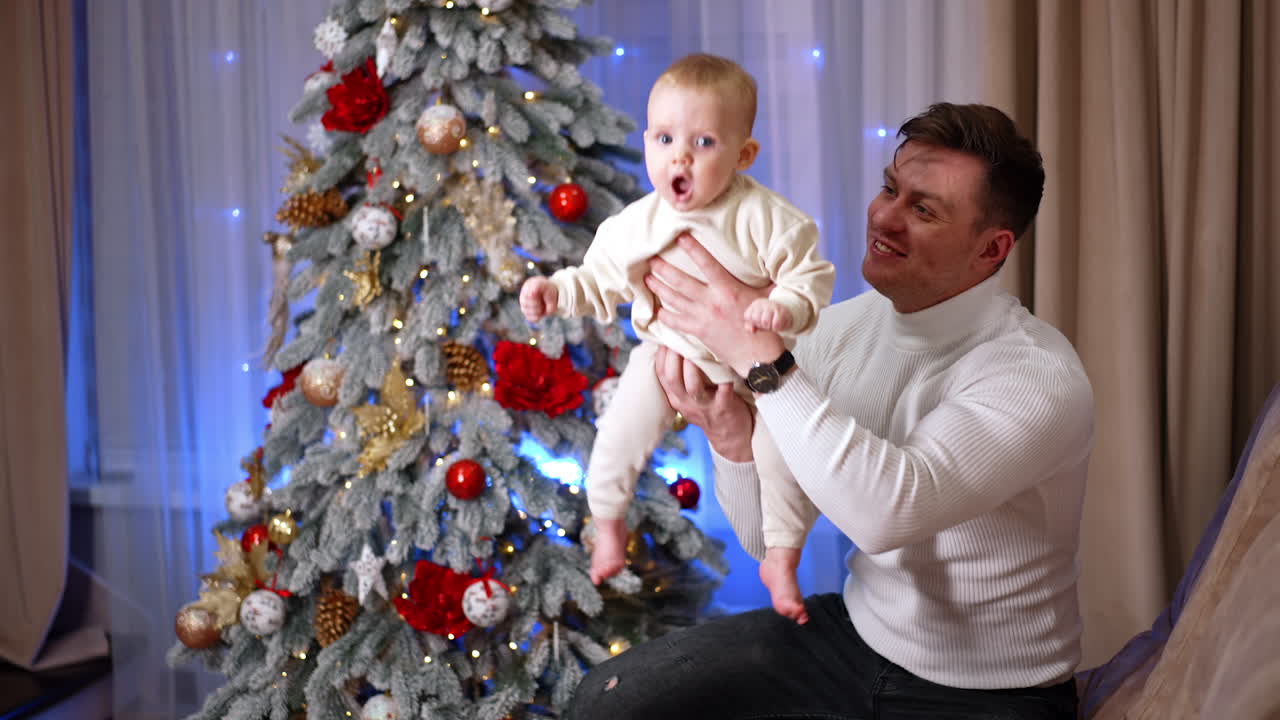 Cheerful Caucasian dad sitting on the sofa plays waving his little baby. Father kisses his son lovingly on the cheek. Christmas tree at backdrop.