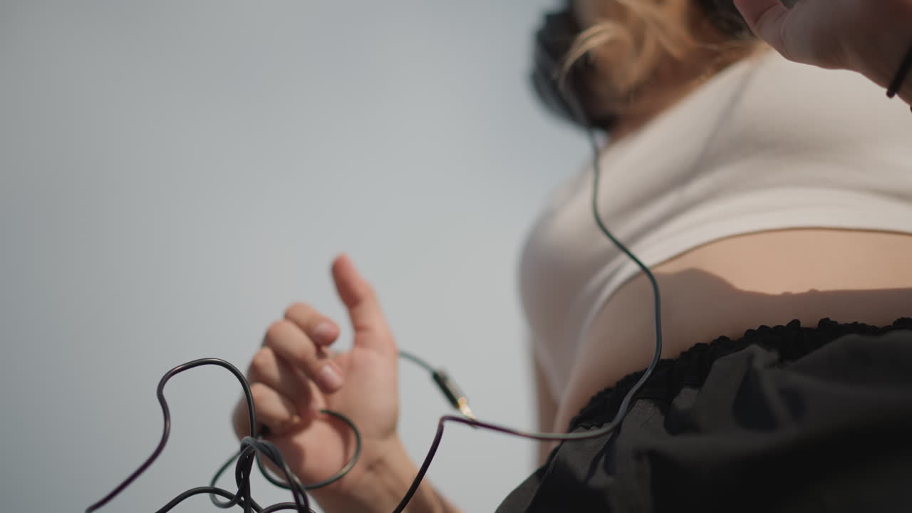 Ung, kaukasisk kvinde justerer ledningsafspiller, solbeskinnet nærbillede af hænder, der løsner ledningen til høretelefonerne og trykker på playlisten på en blå enhed, iført hvid croptop og sorte joggingbukser, diskret hoftebevægelse
