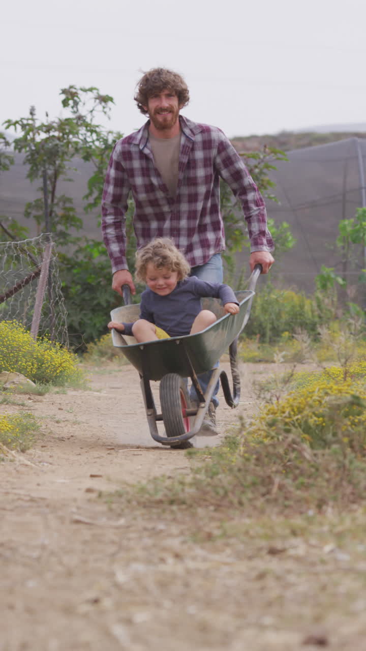 video vertical de un padre caucásico sonriente empujando a su hijo por un camino de campo en una carretilla