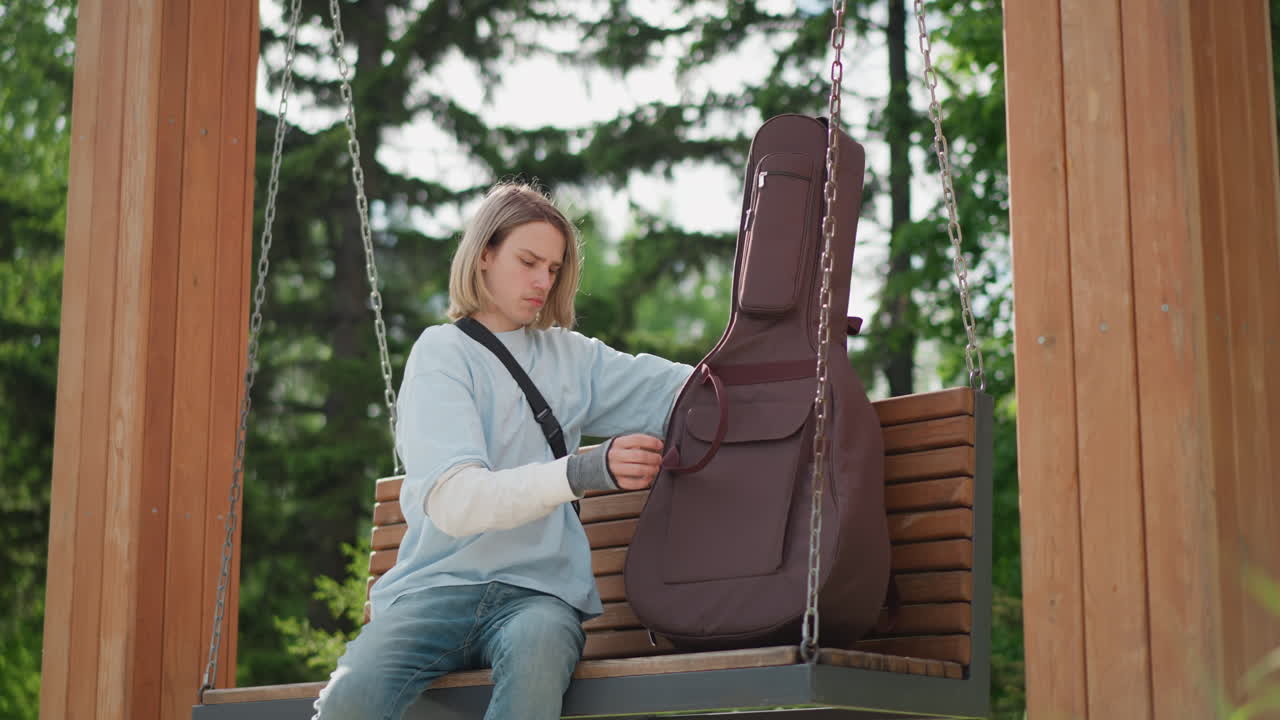 white guitarist packing acoustic guitar on park swing, wooden bench under trees, casual denim outfit, opening padded guitar case, adjusting strap, quiet afternoon sunlight filtering through leaves